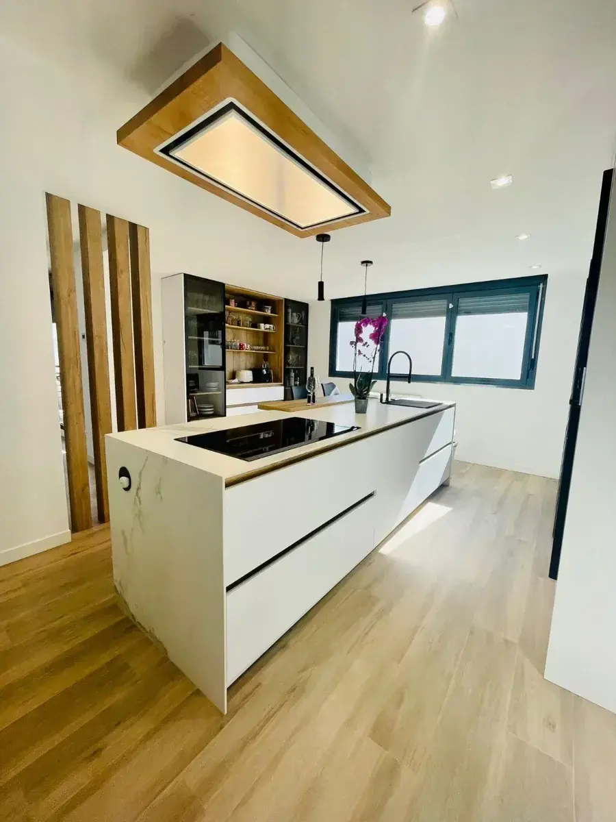 Cocina blanca con isla y campana de madera al techo, vista desde el salón